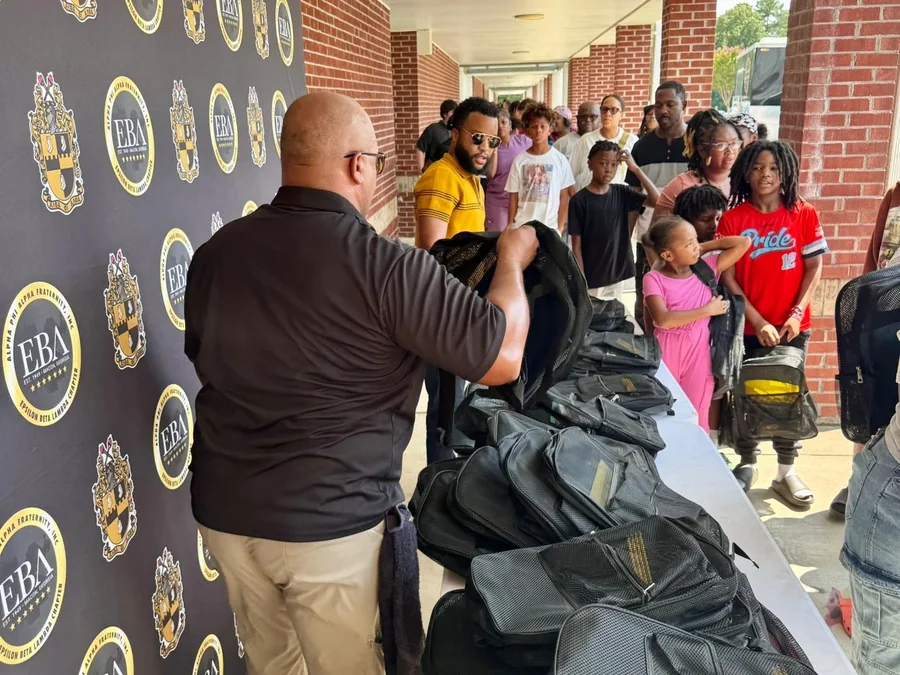 A brother of the chapter with a table of backpacks at a bookbag giveaway, in front of the chapter's banner.