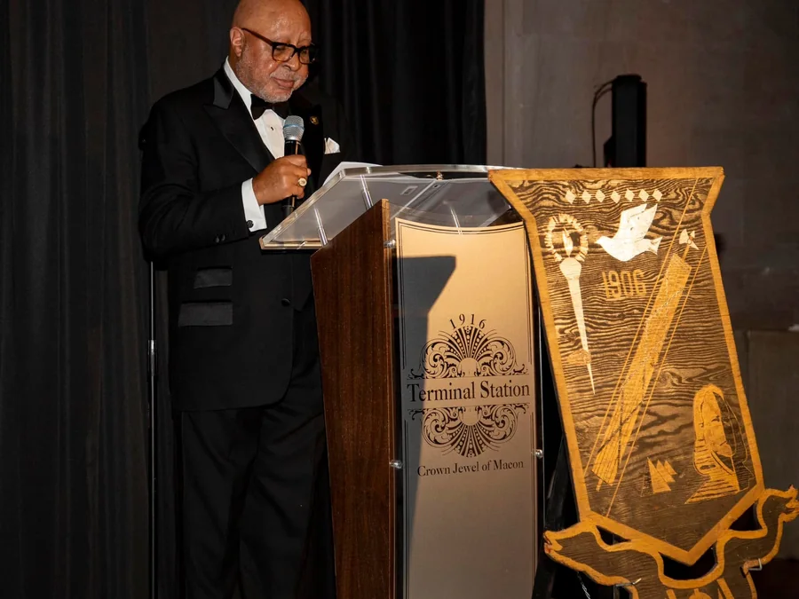 Bro. Chester Wheeler speaking at a podium beneath the Alpha Phi Alpha banner.
