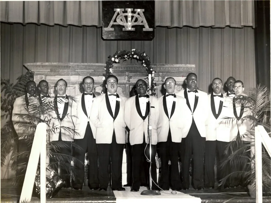 Fourteen brothers in white dinner jackets and black bow ties singing at a microphone on stage, under the Alpha Phi Alpha letters and a flowered arch.