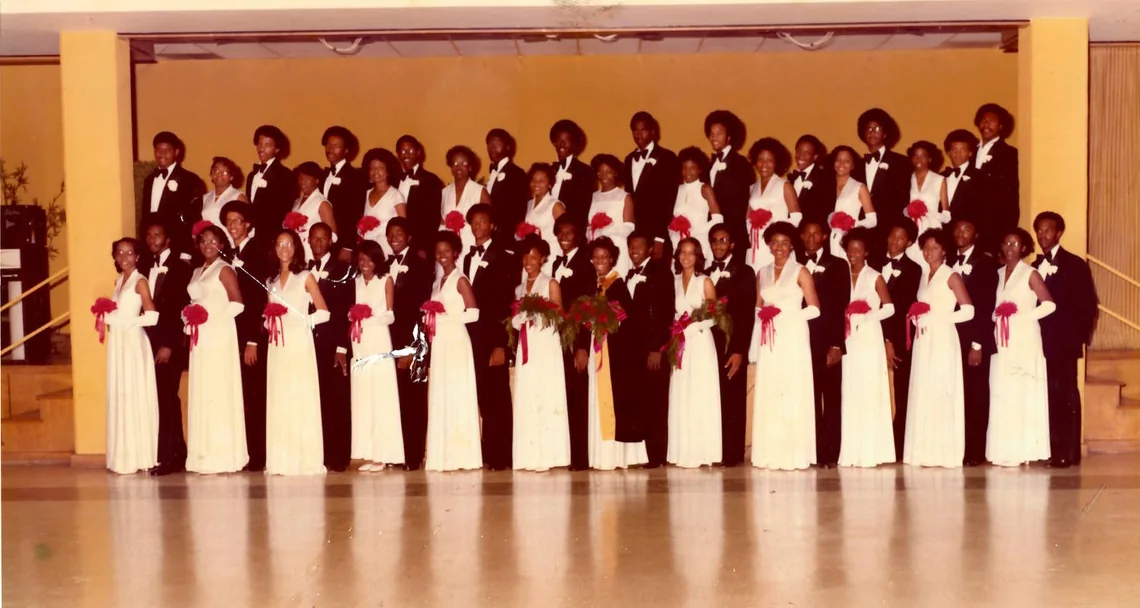 A line of some thirty debutantes in white gowns carrying red bouquets, each beside an escort in black tie, ranked across an auditorium stage.
