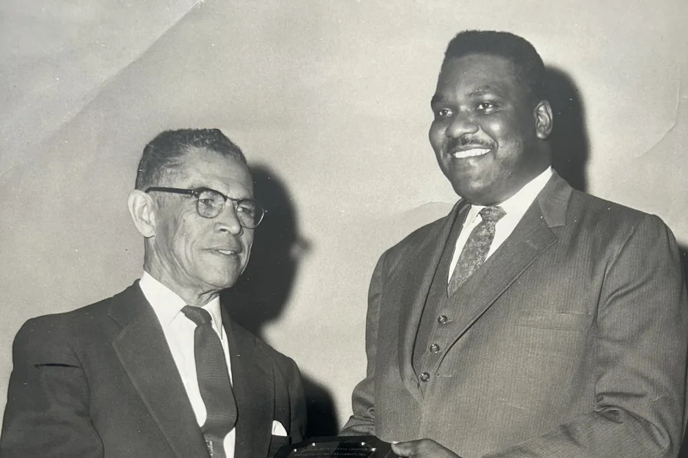 Two men in suits at a presentation, holding an engraved award between them.