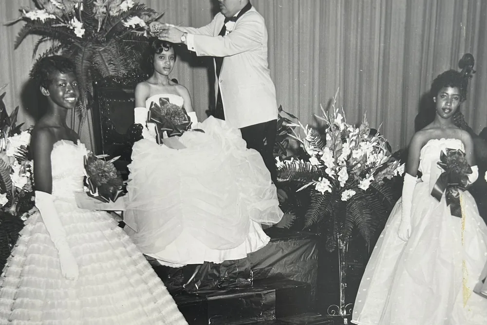 A young woman in a white gown being crowned on a stage banked with flowers, with two more beside her.