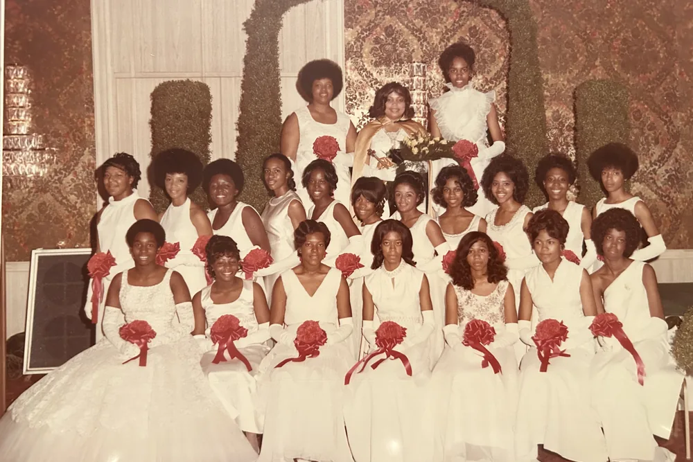 Two rows of young women in white gowns holding red bouquets, on a decorated stage.