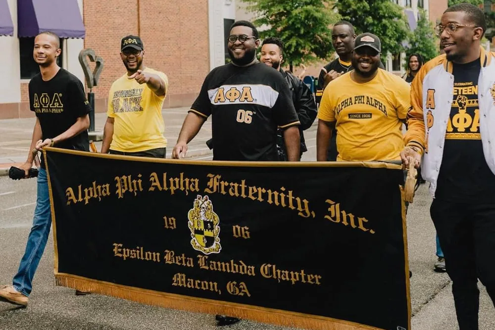 Brothers walking a street behind the chapter banner, which reads Alpha Phi Alpha Fraternity, Inc., 1906, Epsilon Beta Lambda Chapter, Macon, GA.
