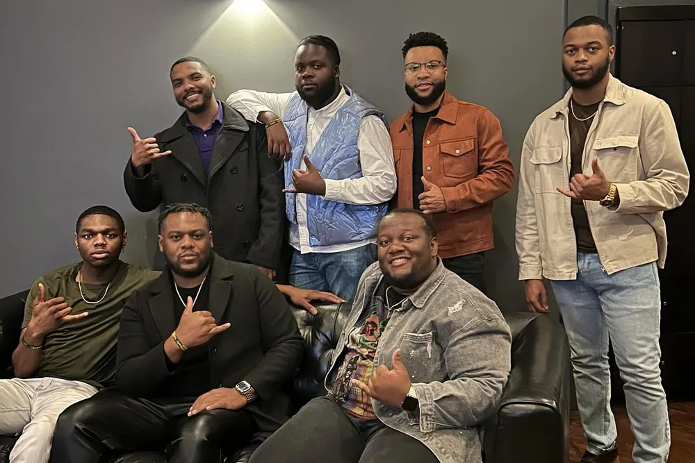 A group of brothers together indoors, several giving the Fraternity's hand sign.